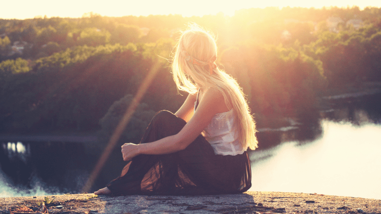 A young woman sitting on top of a mountain lokking over the valley below into the sun.