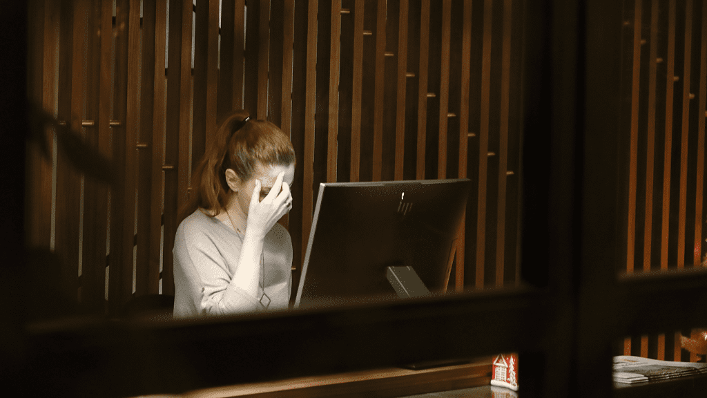 A woman sitting in front of a computer in a dark room with her hand over her face, depressed.