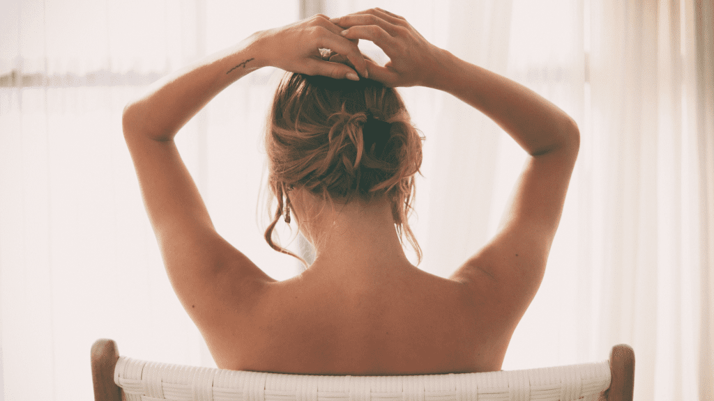A picture of a woman from behind as she sits with bare back in a chair in front of a curtained window.