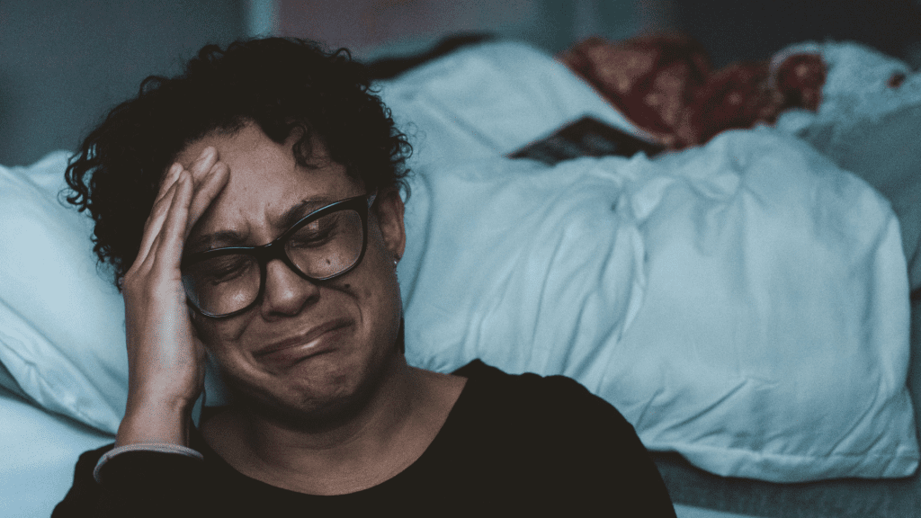 A woman sitting on the floor up against her bed, crying and leaning her head on her hand.