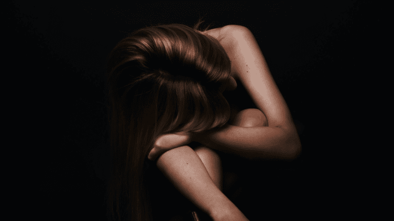 A woman sitting in the dark, head on her knees.