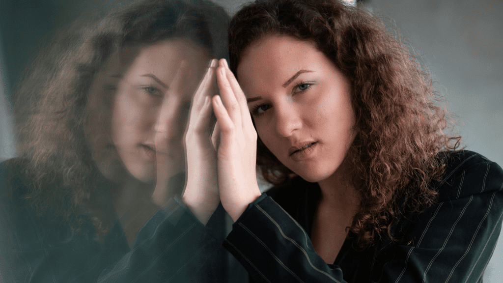 A woman rests against a glass wall, her reflection on the glass, as she stares into the camera.