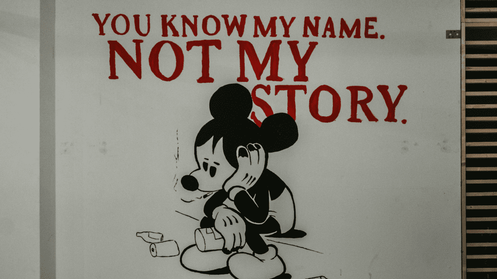 An image of a sad Mickey Mouse sitting on a street corner under the text, "You know my name, not my story."